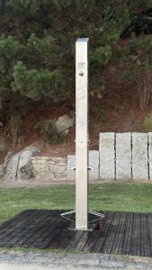 Stainless steel outdoor shower, situated on a black grate on the grass in front of a stone wall and some pine trees.
