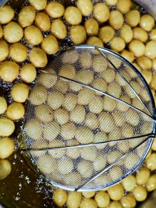 A close-up view of small golden fritters frying in hot oil, with a metal mesh strainer covering the center. The bubbling oil and warm golden tones vividly capture the cooking process. Location: Eranhikkal, Kozhikode, Kerala.