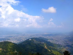 Panoramic view of Pokhara city surrounded by green hills and distant mountains under a partly cloudy sky.