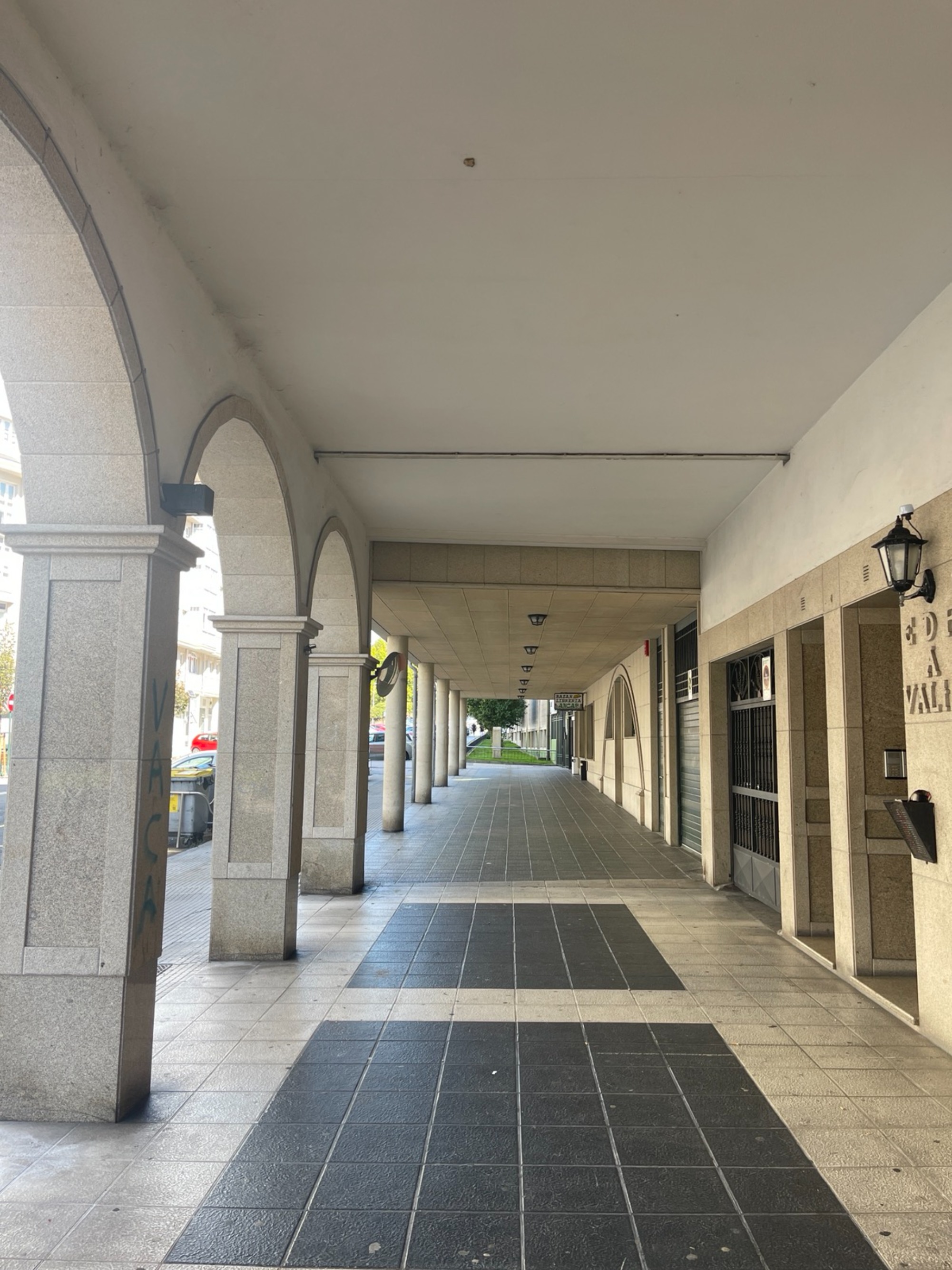 Covered walkway with tall stone arches, tiled floors, closed storefronts, and greenery visible in the distance.