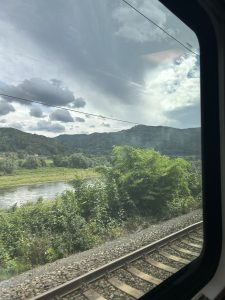 Panoramic view from a train of a river and mountains under a cloudy sky.