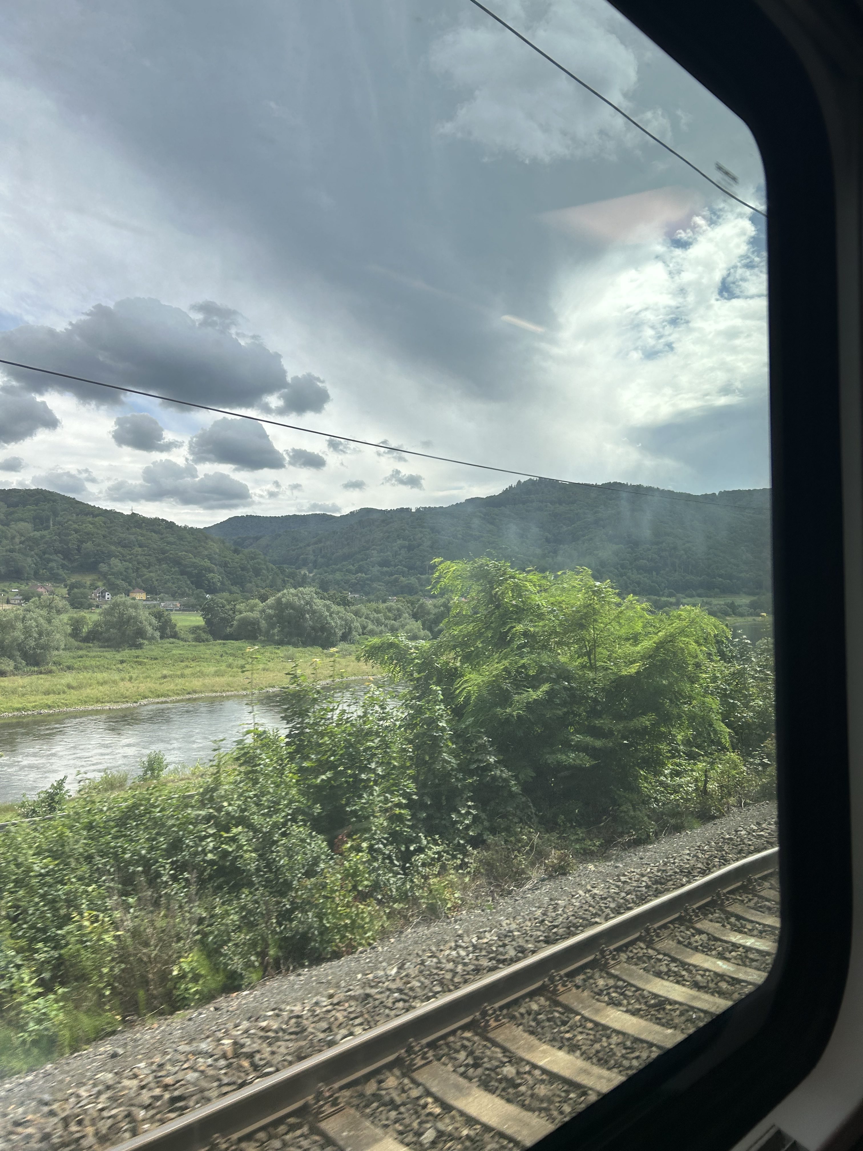Panoramic view from a train of a river and mountains under a cloudy sky.