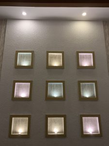 A textured light wall with nine square recessed niches in a 3×3 grid. Each niche has a warm yellow-brown frame and a soft glowing light at its base.
