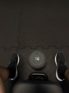 Top-down view of black and blue sneakers and a black 10 kg dumbbell on a black rubber gym mat.