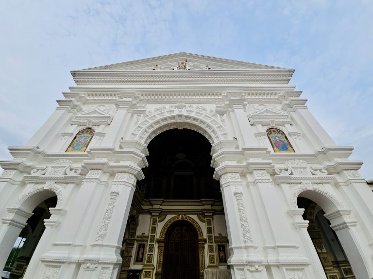 A close-up view of the ornate white façade of a historic church, featuring decorative arches, columns, stained-glass windows, and intricate relief work. Location: Ramapuram, Kottayam, Kerala