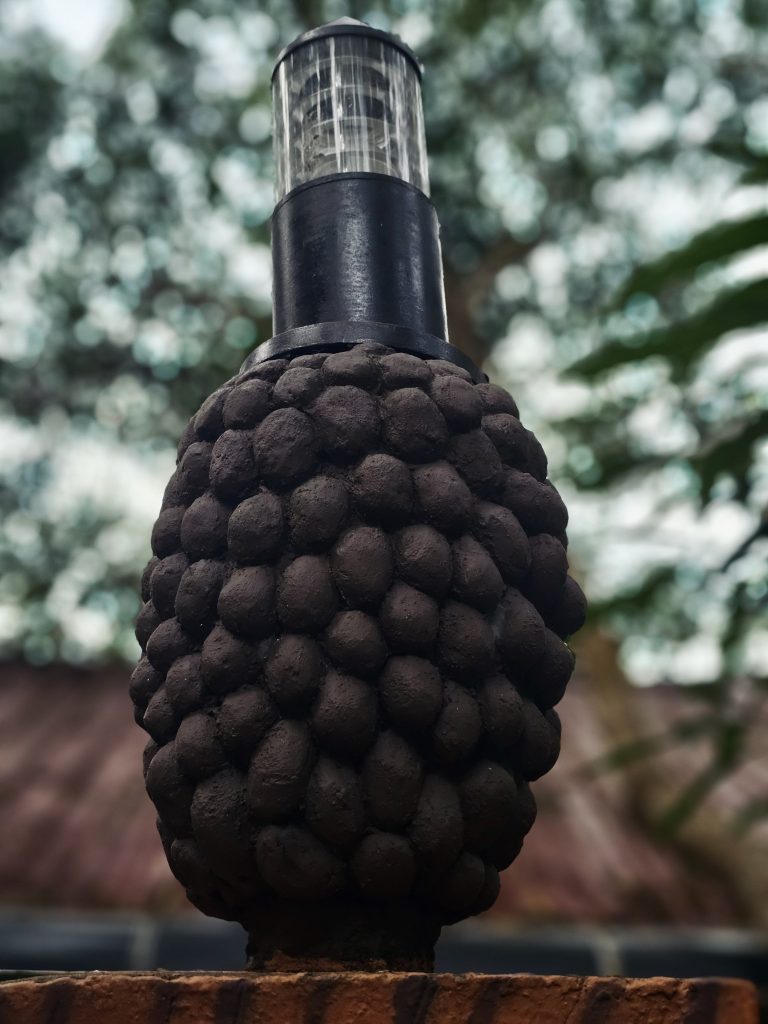 A distinctive textured outdoor lamp (Mulberry style) with a rounded, layered surface is photographed against a softly blurred natural background. Location: Ayamkudy, Kottayam, Kerala.