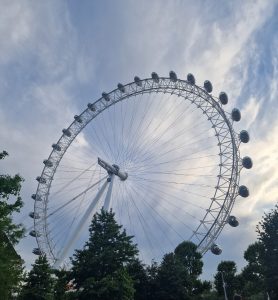 Low-angle view of the large white wheel structure of the London Eye rising above dark green summer trees under a cloudy sky.