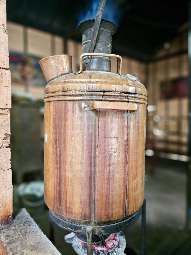 A traditional copper water heater, commonly known as a bamba or South Indian samovar, used to heat water, sits over glowing charcoal, highlighting its aged metal fittings and rustic construction in detail. The shallow depth of field isolates the object nicely. Location: Ayamkudy, Kottayam, Kerala.