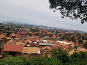 A panoramic view of a densely populated area featuring numerous houses with brown and red metal roofs. The landscape includes patches of greenery, trees, and a distant hilly horizon. 