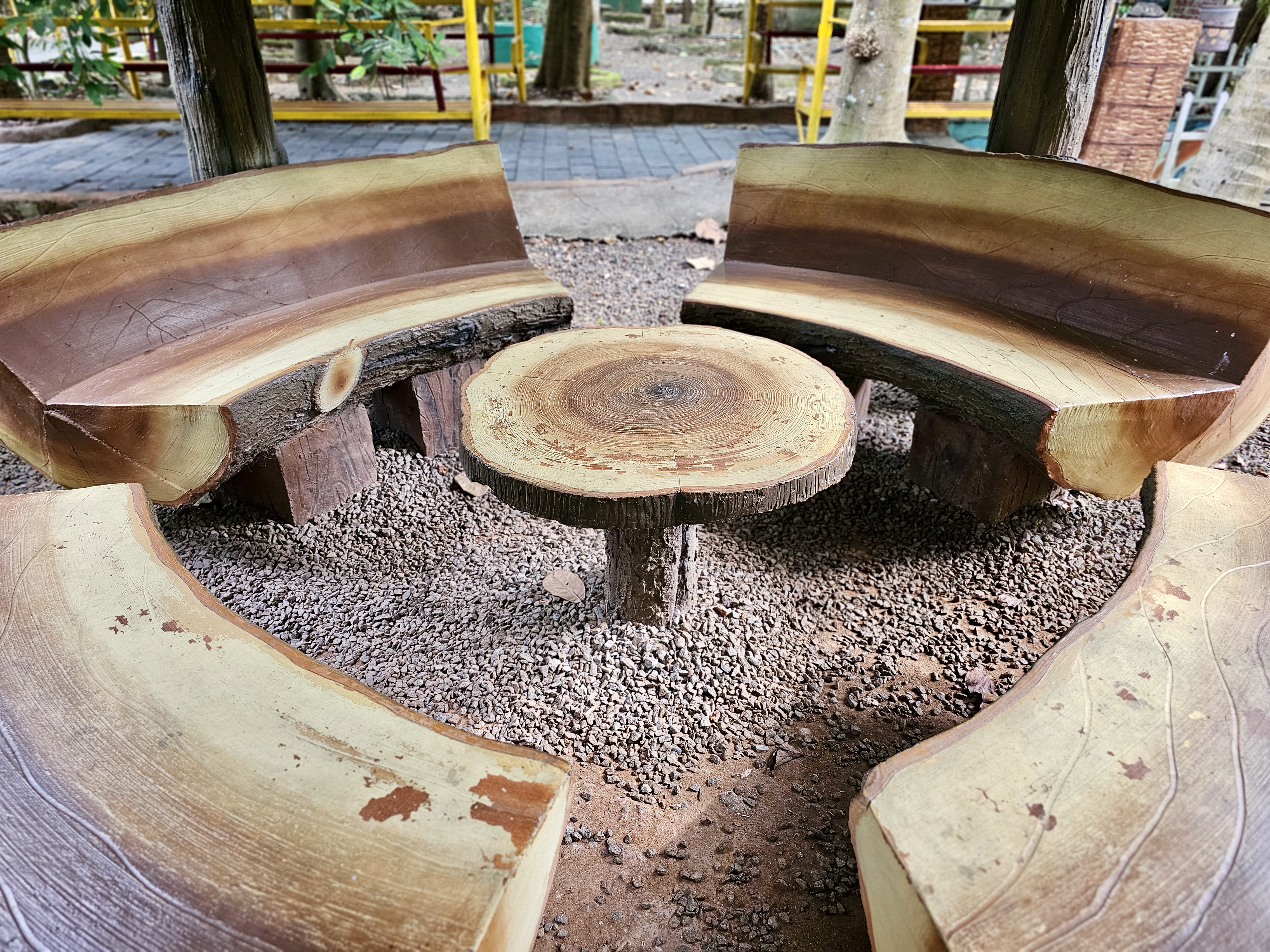 Rustic concrete benches and a round tree-stump table, both designed with wooden patterns, are arranged in an outdoor seating area. The natural wood-like textures and circular arrangement create an interesting visual. Location: Ayamkudy, Kottayam, Kerala.