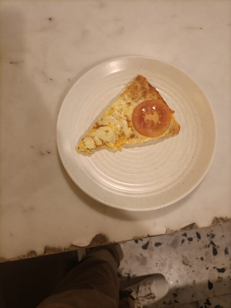 A slice of pizza sits on a round, light-colored plate with a textured surface.
