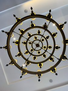 Upward view of an ornate chandelier with concentric bronze rings and black-and-gold lights, forming a dramatic geometric pattern.