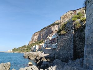 A wall and a couple of houses over rocks on the water along a clear sky behind a field.