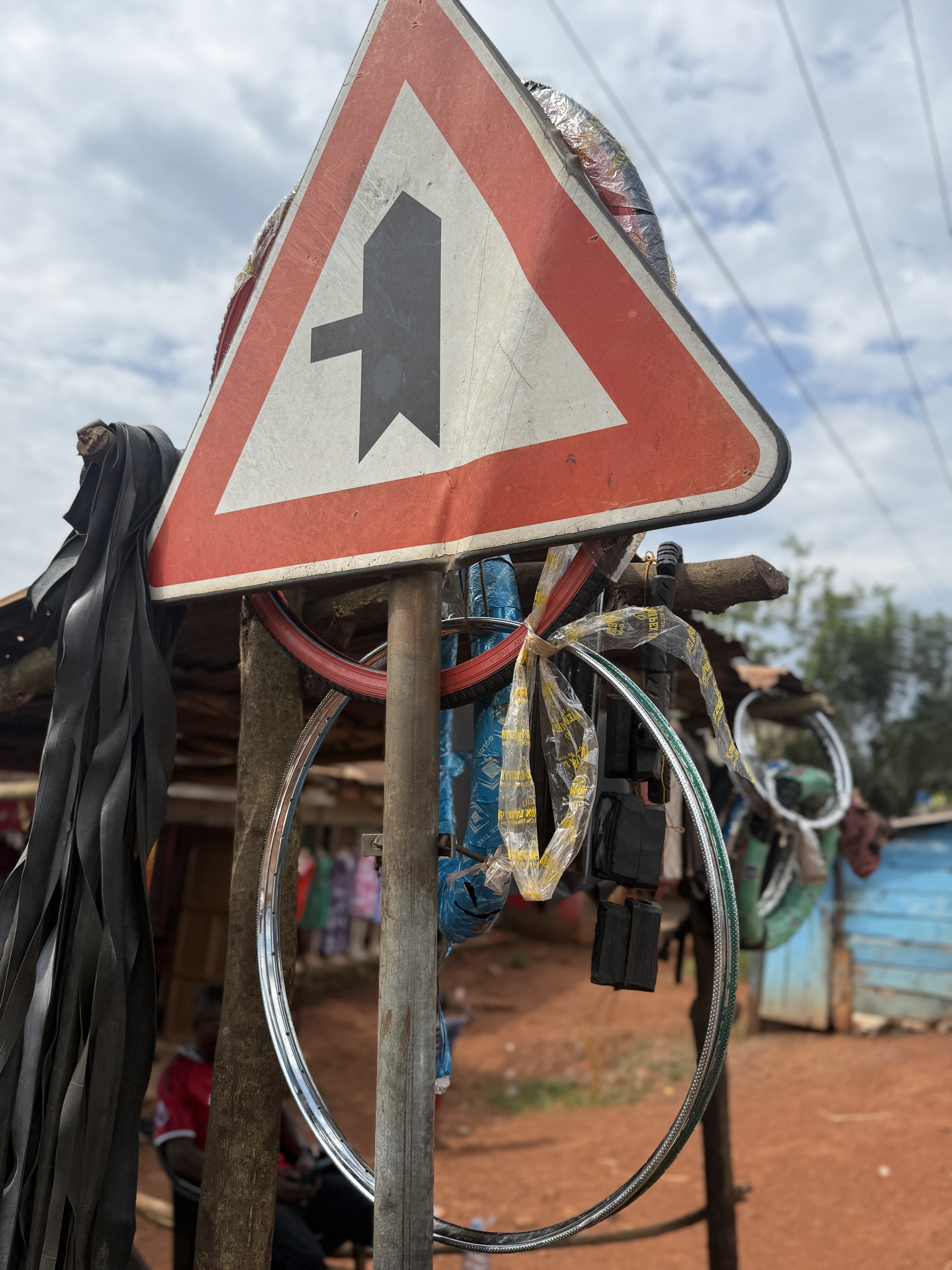 Triangular speed bump warning sign on a metal post, surrounded by wires and straps, with roadside structures and reddish dirt in the background.