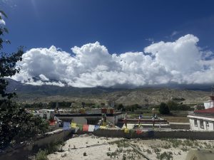 An elevated view of traditional flat-roofed stone buildings decorated with colorful Buddhist prayer flags stretched across lines. Dense, dramatic white clouds blanket rolling dry hills under a vivid blue sky.