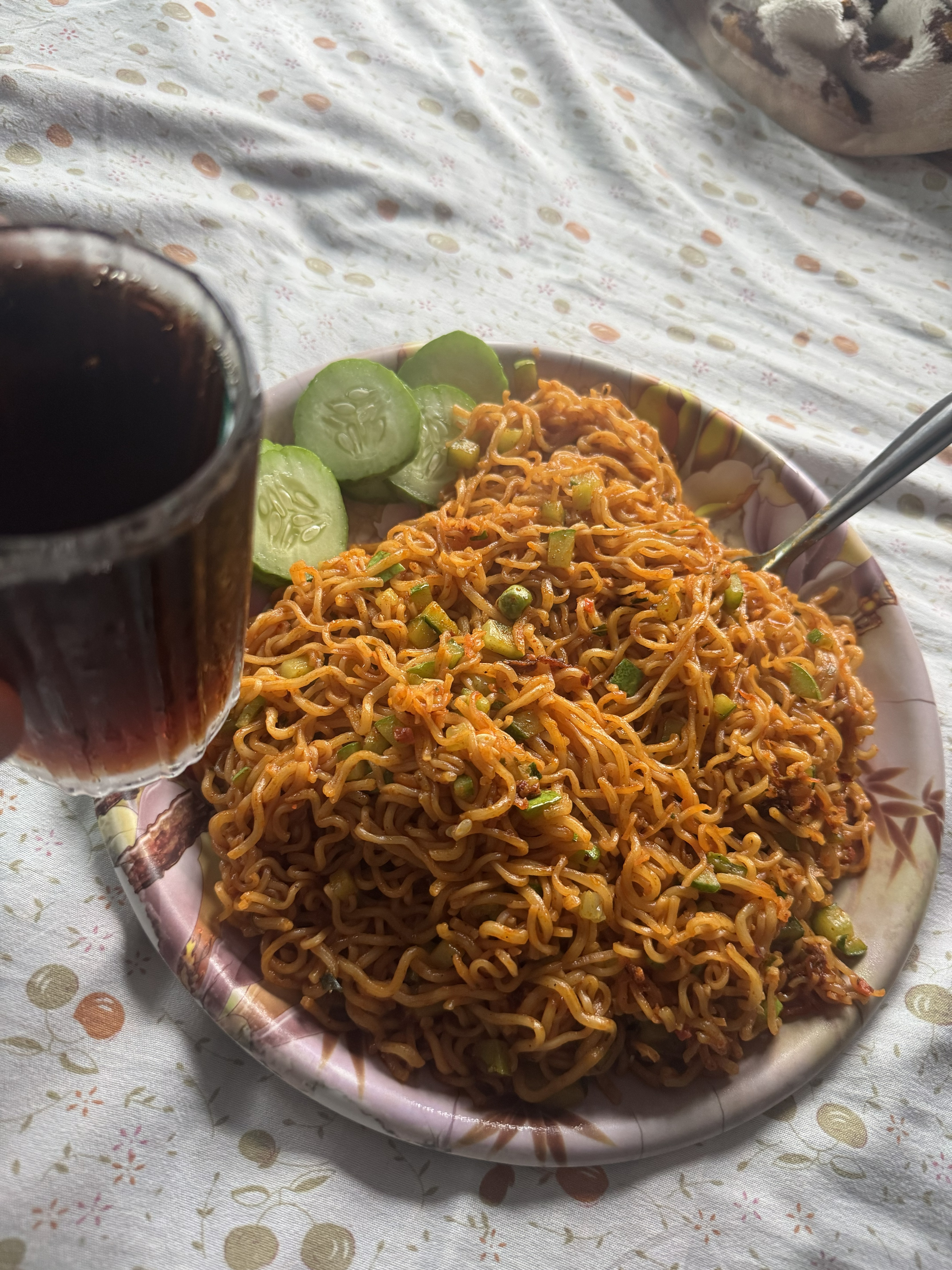 A plate filled with stir-fried spicy noodles mixed with chopped veggies and served with cucumber slices on the side, accompanied by a glass of dark soda resting on a patterned bedsheet.