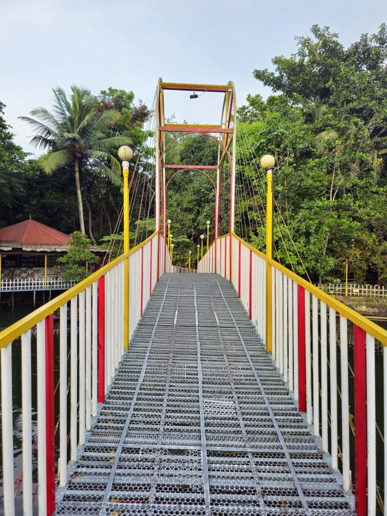 A colorful pedestrian suspension bridge surrounded by lush tropical vegetation, with a calm body of water visible below. The symmetrical composition and the bridge's repeating lines create a strong sense of depth. Location: Ayamkudy, Kottayam, Kerala.