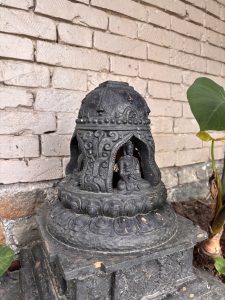 Small black stupa sculpture with intricate patterns and a seated figure, placed against a light brick wall with a few green leaves nearby.
