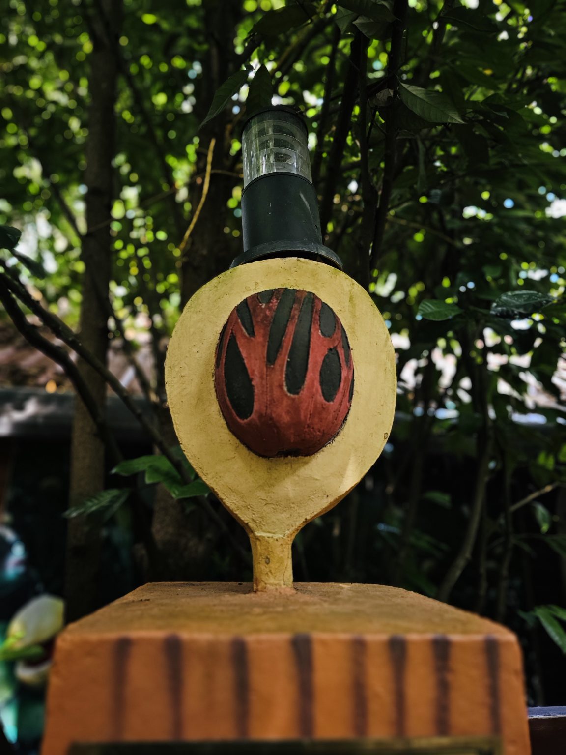 A decorative outdoor lamp designed with a rustic, earthy appearance, featuring a painted red-and-black motif and surrounded by dense tropical greenery. Location: Ayamkudy, Kottayam, Kerala.