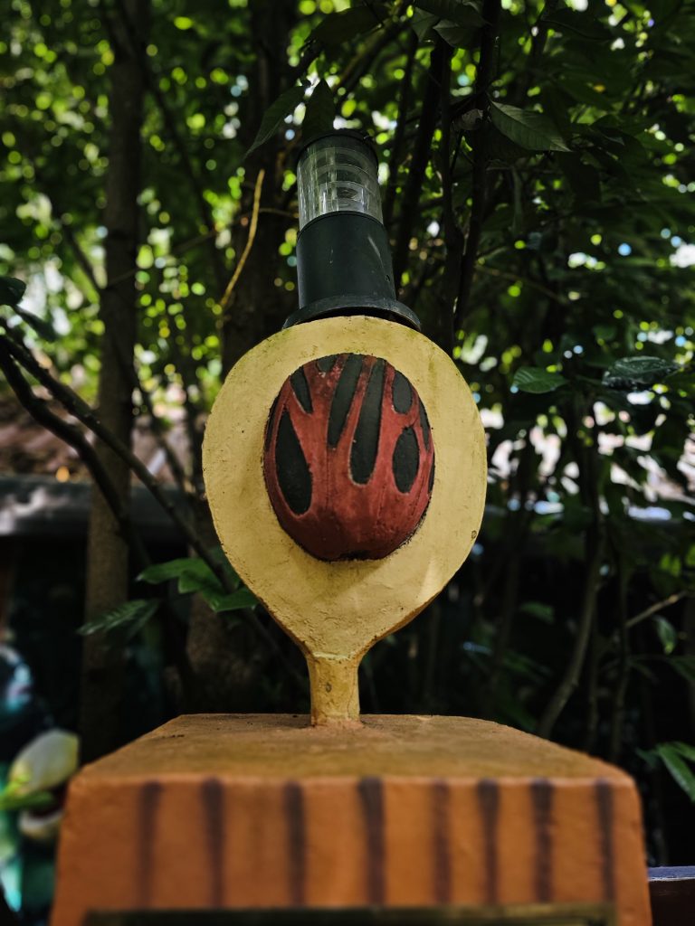 A decorative outdoor lamp designed with a rustic, earthy appearance, featuring a painted red-and-black motif and surrounded by dense tropical greenery. Location: Ayamkudy, Kottayam, Kerala.