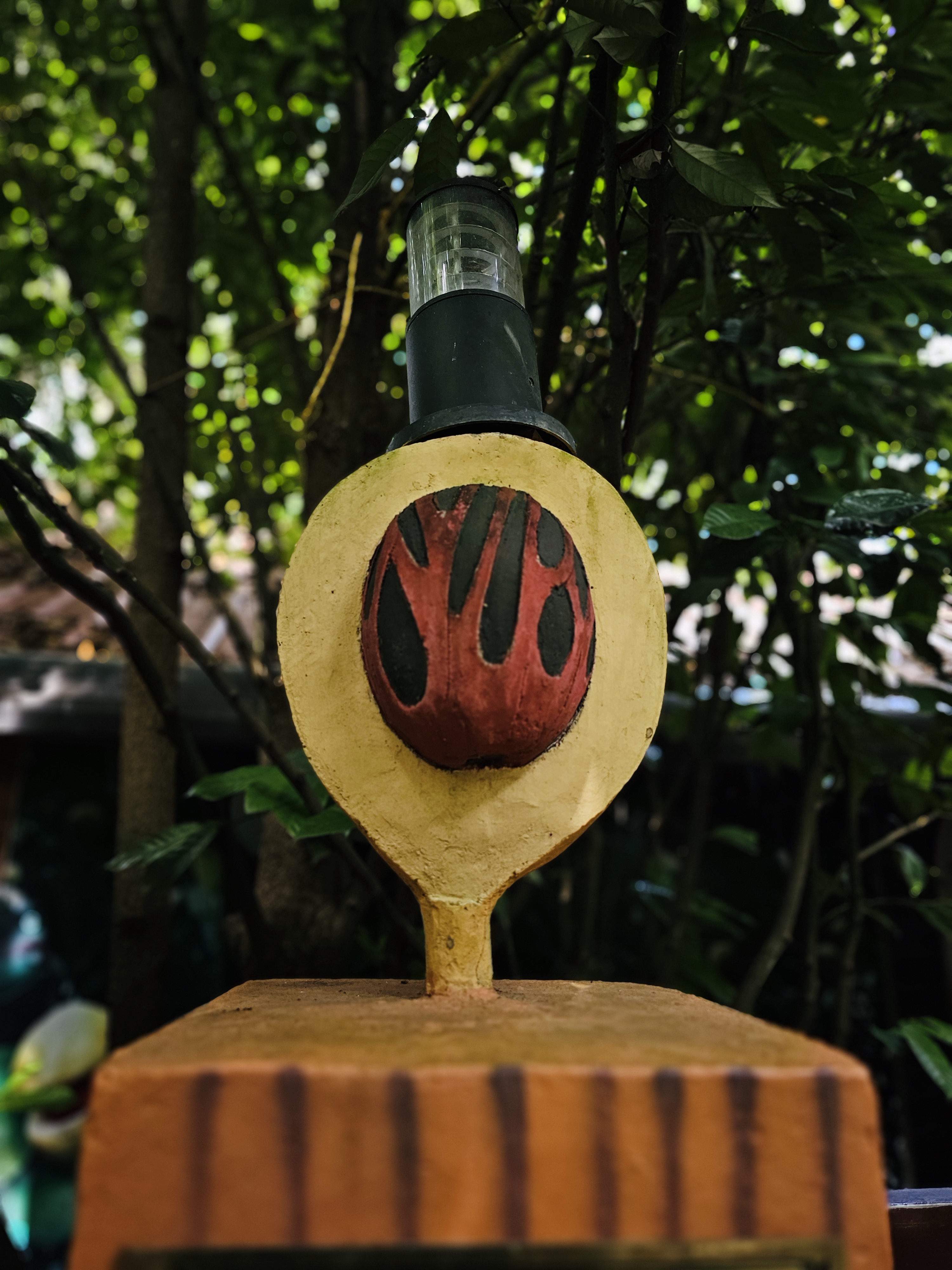 A decorative outdoor lamp designed with a rustic, earthy appearance, featuring a painted red-and-black motif and surrounded by dense tropical greenery. Location: Ayamkudy, Kottayam, Kerala.