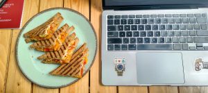 Grilled vegetable sandwiches served on a ceramic plate beside a laptop on a wooden table.
