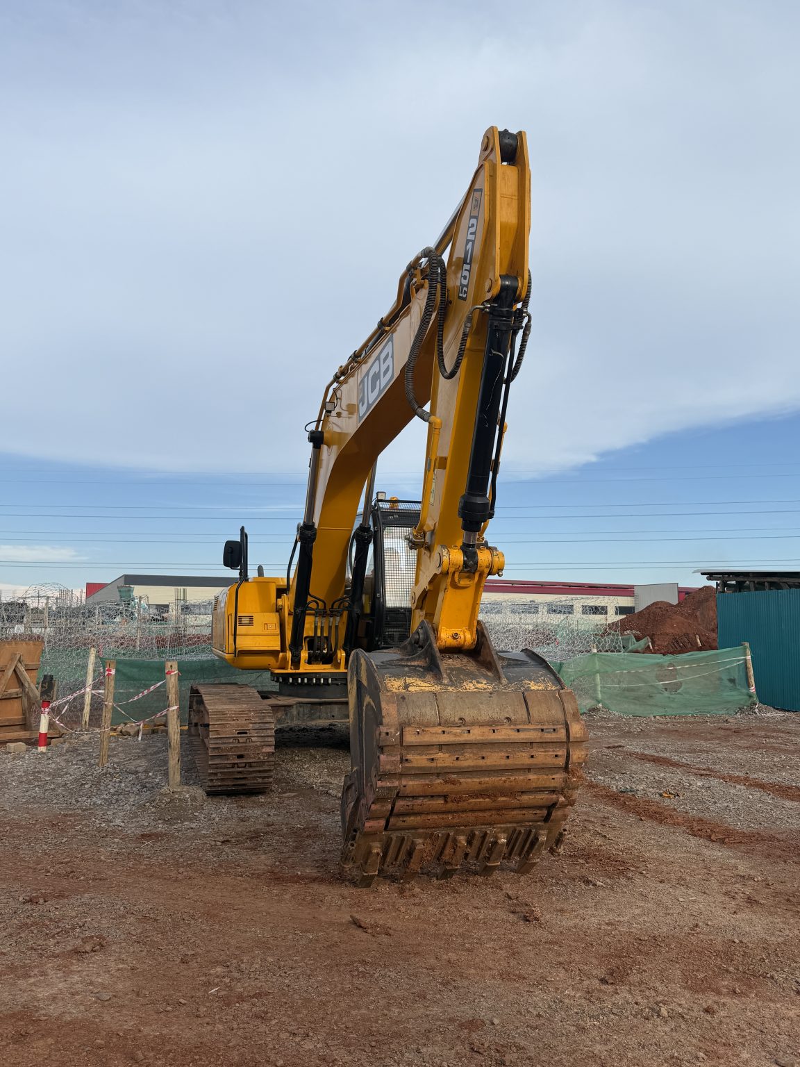 A yellow excavator with its bucket resting on the ground sits on a dirt construction site, with fencing, piles of soil, and buildings visible in the background under a partly cloudy sky.