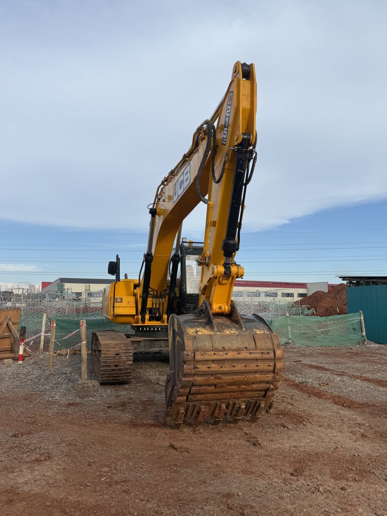 A yellow excavator with its bucket resting on the ground sits on a dirt construction site, with fencing, piles of soil, and buildings visible in the background under a partly cloudy sky.