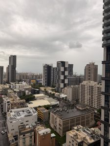 High-angle view over Kaohsiung, Taiwan on an overcast day, with modern high-rise towers, flat rooftops, and a rooftop parking lot, and port cranes and a Ferris wheel on the hazy horizon.
