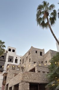 Old multi-storey coral-stone house in historic Al-Balad, Jeddah, strung with warm glowing bulbs, next to white buildings with dark wooden balconies and a tall palm tree leaning across a clear sky.
