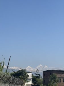 A clear view of snow-capped Himalayan mountain peaks visible above tree lines, brick buildings, and razor wire fencing under a bright blue sky