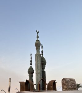 Bronze sculpture of three ornate minarets topped with crescents, rising from curved rust-coloured arches on a low stone plinth, beside a crumbling coral-stone column in Al-Balad, Jeddah, against a pale evening sky.
