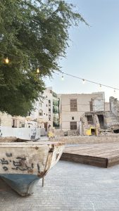 Weathered wooden boat with hand-painted Arabic lettering resting on a paved courtyard in Al-Balad, Jeddah, under a leafy tree and a string of lights, with a ruined stone house and historic buildings behind.
