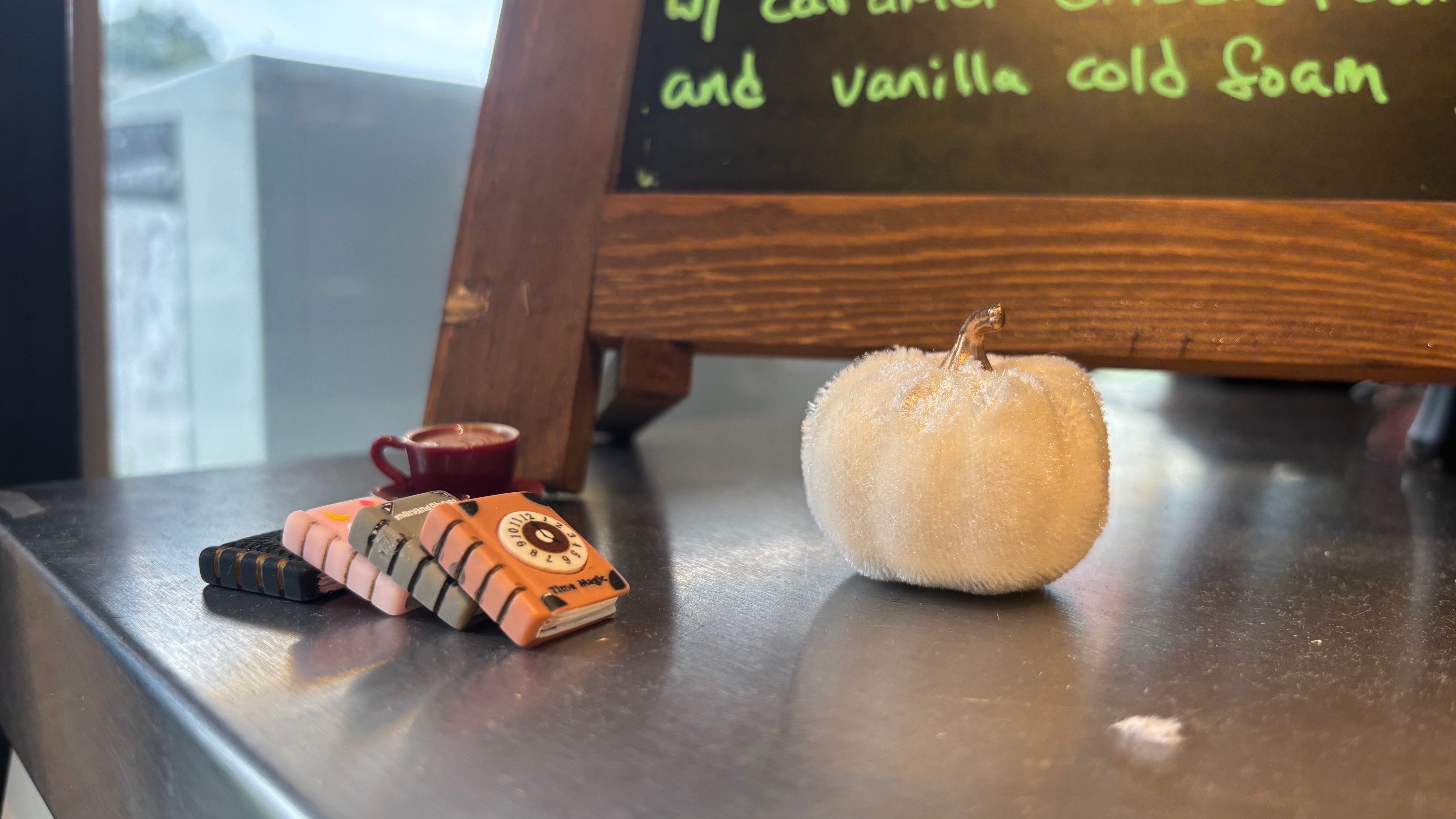Close-up of the bottom edge of a coffee shop sign. Right in front of it are miniature Halloween books and a miniature pumpkin.