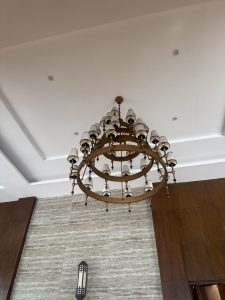 A large multi-tiered brass ring chandelier featuring numerous small lampshades, suspended from a white ceiling above textured stone and wood wall cladding.