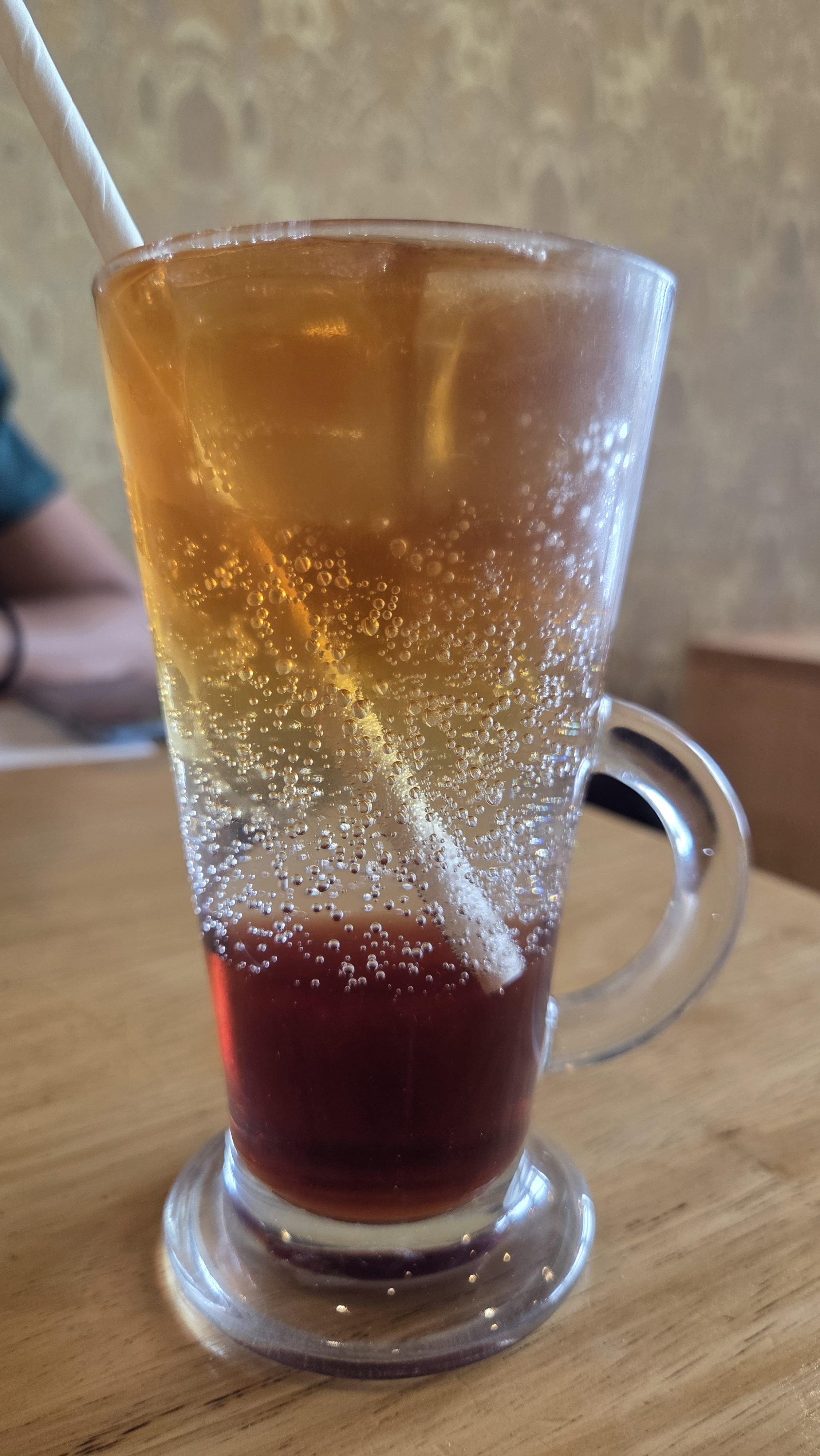 A tall glass of Red Autom, a sparkling golden-orange drink with a deep red layer at the bottom, filled with bubbles and served with a white straw on a wooden table.