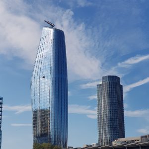Wide-angle view looking up at the curved glass facade of the One Blackfriars tower rising into a blue sky with soft clouds.