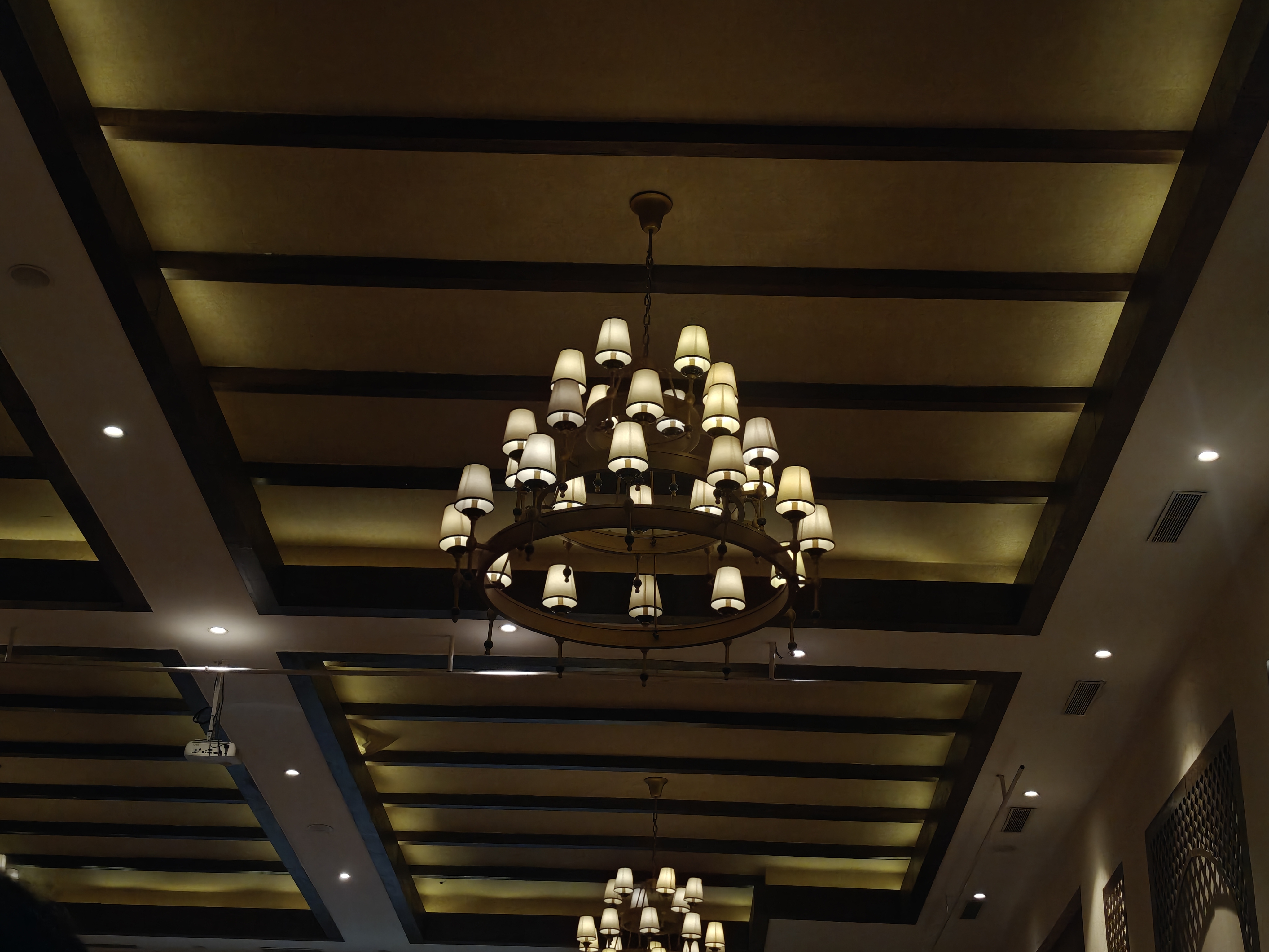Large circular chandelier with cream lampshades hanging from a wooden grid ceiling in a warmly lit interior.
