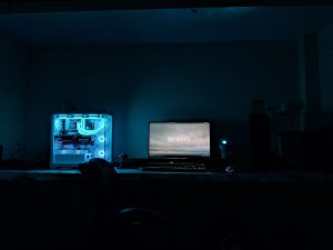 The image shows a dimly lit workspace featuring a gaming computer setup. On the left, there’s a large, transparent computer case with glowing blue LED lights illuminating the interior components. To the right, a monitor displays a screen with a blurred background and the text "52 BARS," set against a dark theme. In front of the monitor, a black mechanical keyboard and a colorful gaming mouse are visible. A small, round blue light source sits next to the monitor. The overall ambiance is cool and tech-oriented, suggesting a modern gaming environment.
