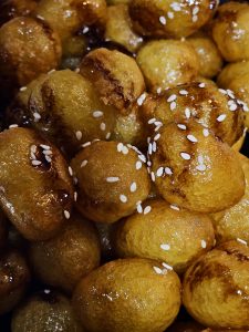 A close-up of golden, syrup-coated sweet fritters garnished with white sesame seeds. The glossy surface, rich golden color, and tight composition make this an appetizing food photograph. Location: Eranhikkal, Kozhikode, Kerala