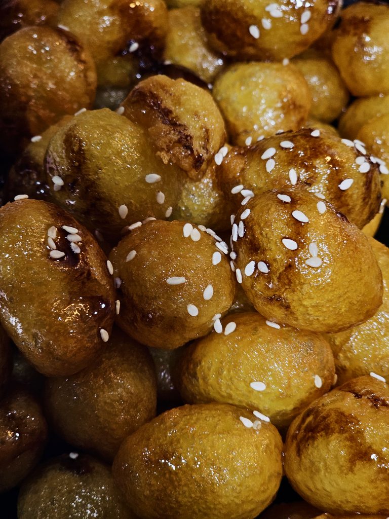 A close-up of golden, syrup-coated sweet fritters garnished with white sesame seeds. The glossy surface, rich golden color, and tight composition make this an appetizing food photograph. Location: Eranhikkal, Kozhikode, Kerala