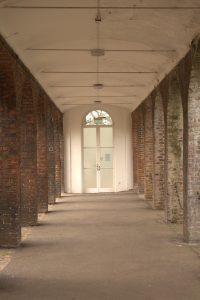Straight-on view through an outdoor red brick vaulted archway corridor looking toward an arched white glass door at the end.