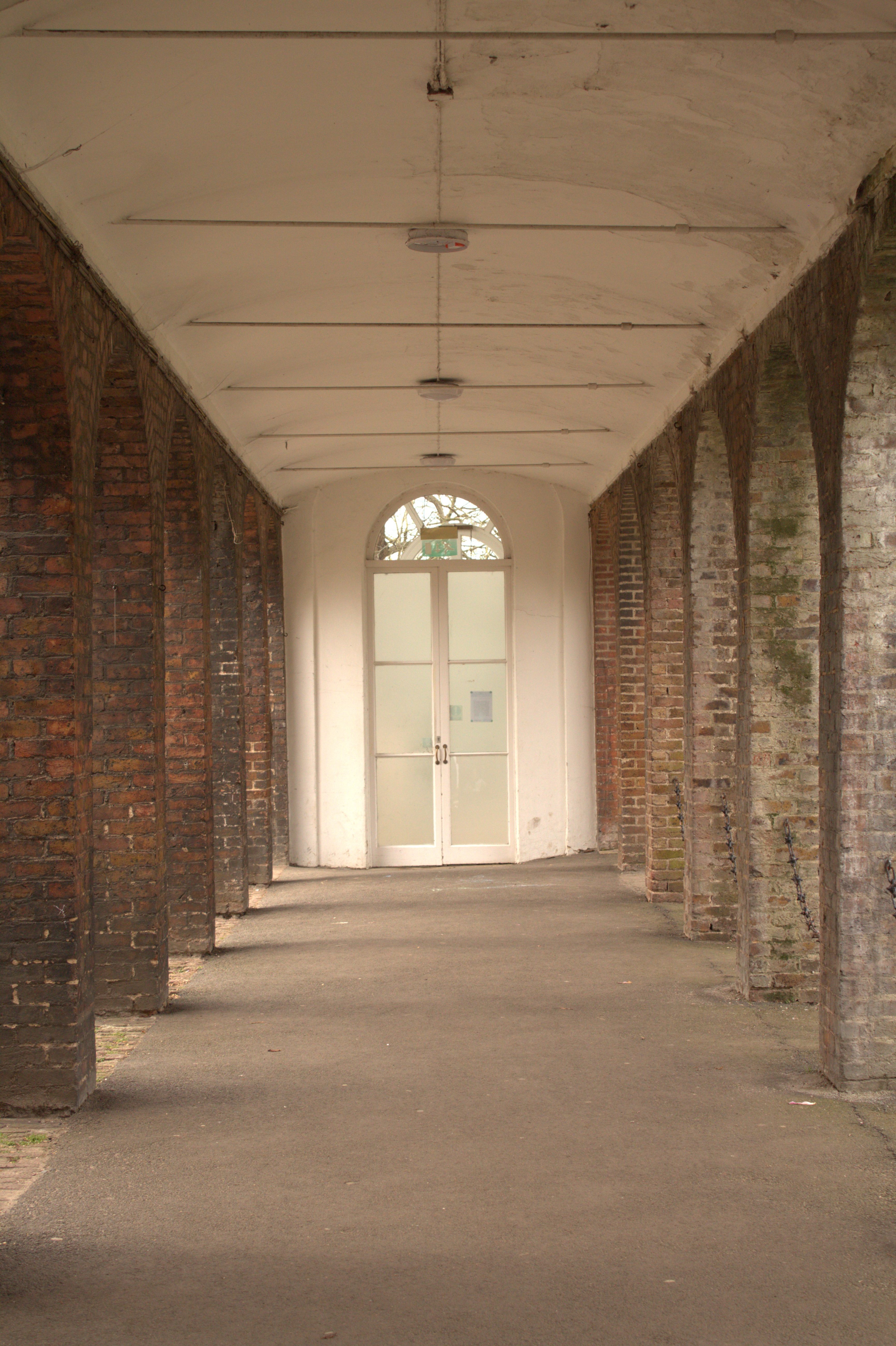Straight-on view through an outdoor red brick vaulted archway corridor looking toward an arched white glass door at the end.