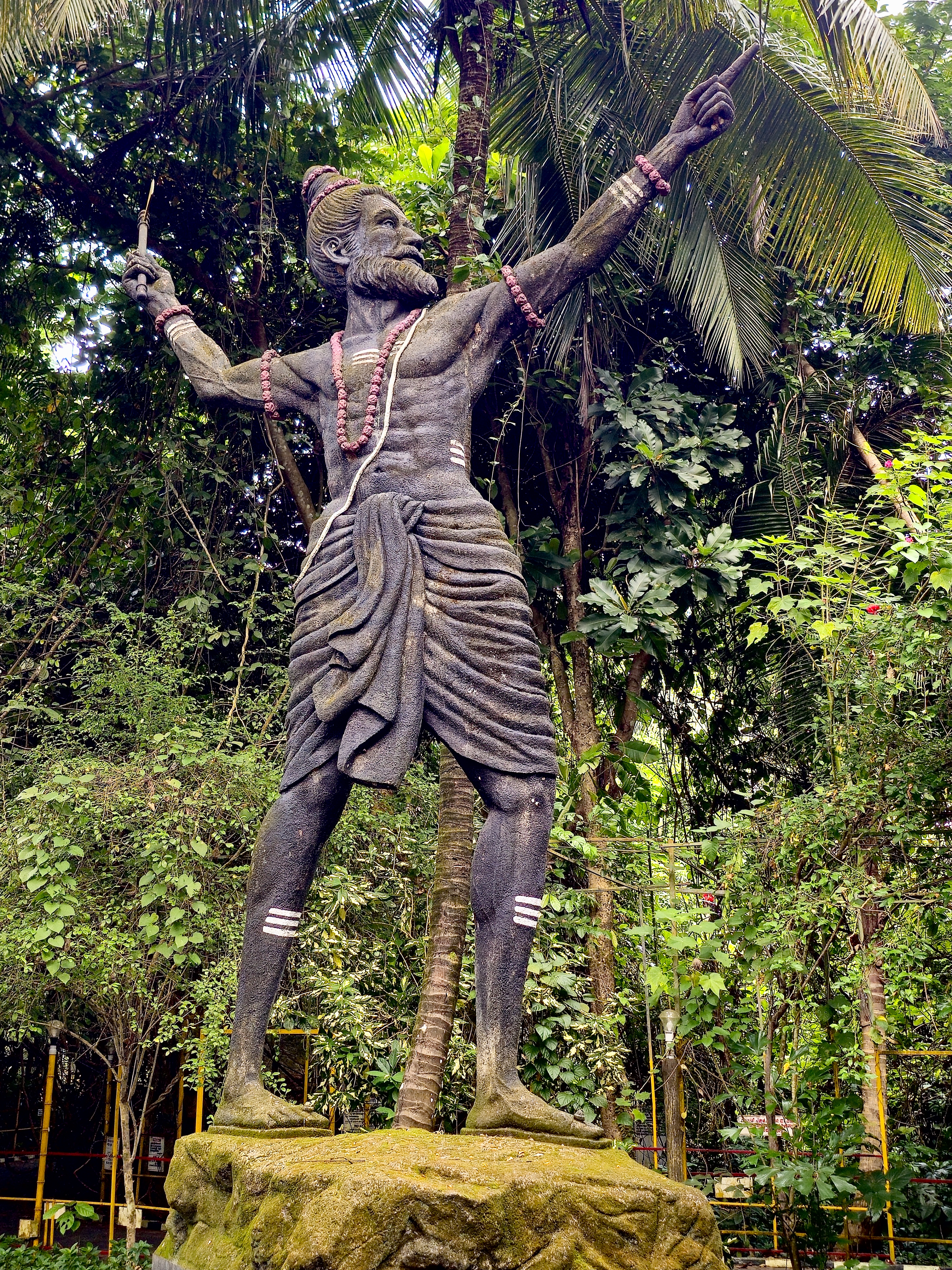 A detailed outdoor statue of a traditional bearded figure stands among coconut palms and dense tropical vegetation. The upward-reaching pose and natural surroundings create a strong cultural and environmental composition. Location: Ayamkudy, Kottayam, Kerala.
