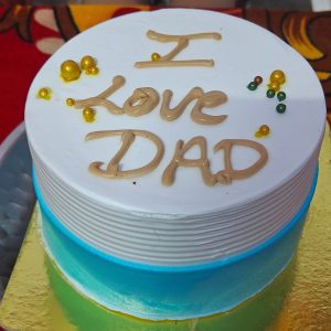 Round birthday cake with white and blue frosting, “I Love Dad” written on top, and decorated with golden and green sugar pearls.
