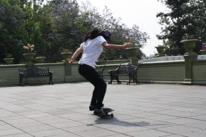 A person wearing a white t-shirt, dark pants, and a hat skateboarding on a paved plaza, captured mid-motion near park benches and green foliage.