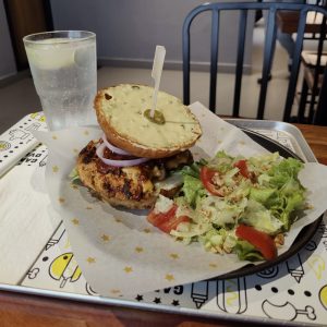 Grilled chicken burger topped with melted cheese, barbecue sauce, and sliced red onion, served with a fresh lettuce and tomato salad on a tray, alongside a glass of sparkling water.