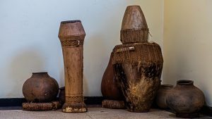 Collection of traditional Ugandan handmade wooden drums and clay pots displayed against a plain light-colored wall on a tiled floor.