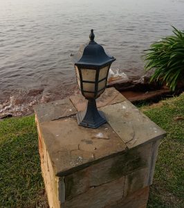 A vintage black metal lantern-style outdoor lamp mounted on a stone pillar along a grassy part of the Lake Victoria shoreline with gentle water waves visible.