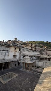 Historic white stone palace complex with ornate domes, arches, balconies, and courtyards, set against a green hillside beneath a clear blue sky.
